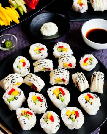 Large black round plate with a lot of white sushi rolls with colorful filling sprinkled with tiny black sesame seeds. Condiment bowls are around it.
