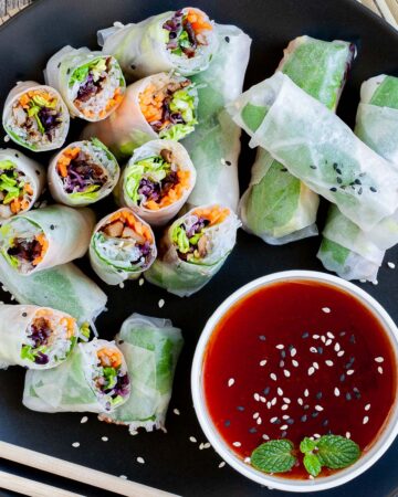 Opaque rice paper rolls filled with carrots, purple sprouts, thin glass. noodles and brown mushrooms are served on a large black plate. Several rolls are cut half so you can see the filling. Red sauce in a small white bowl is served next to it.