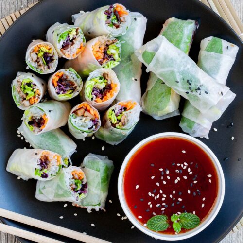 Opaque rice paper rolls filled with carrots, purple sprouts, thin glass. noodles and brown mushrooms are served on a large black plate. Several rolls are cut half so you can see the filling. Red sauce in a small white bowl is served next to it.