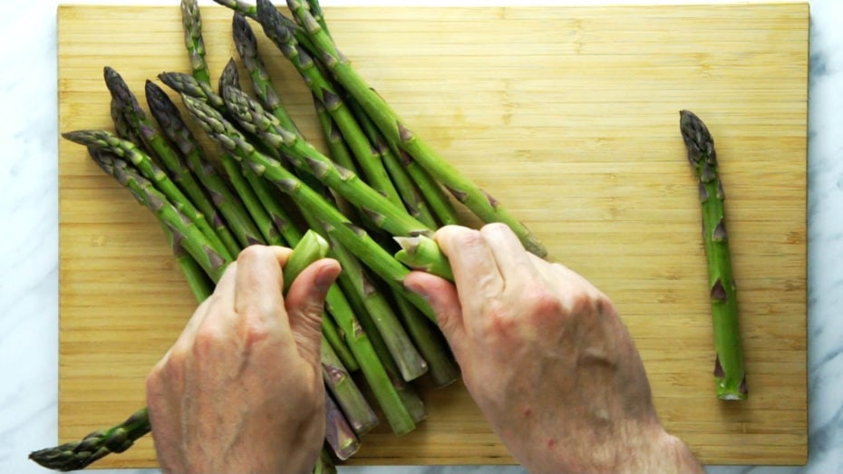 Best spring side dish with asparagus and mushrooms roasted in the oven and ready in under 20 minutes. Easy, delicious and suitable for all diets (vegan, paleo, gluten-free, whole30, keto). #roastedasparagus #asparagusandmushrooms #roastedmushrooms #asparausrecipe #roastedveggies Close-up of how to snap asparagus stalk with hands above a wooden board full of fresh asparagus