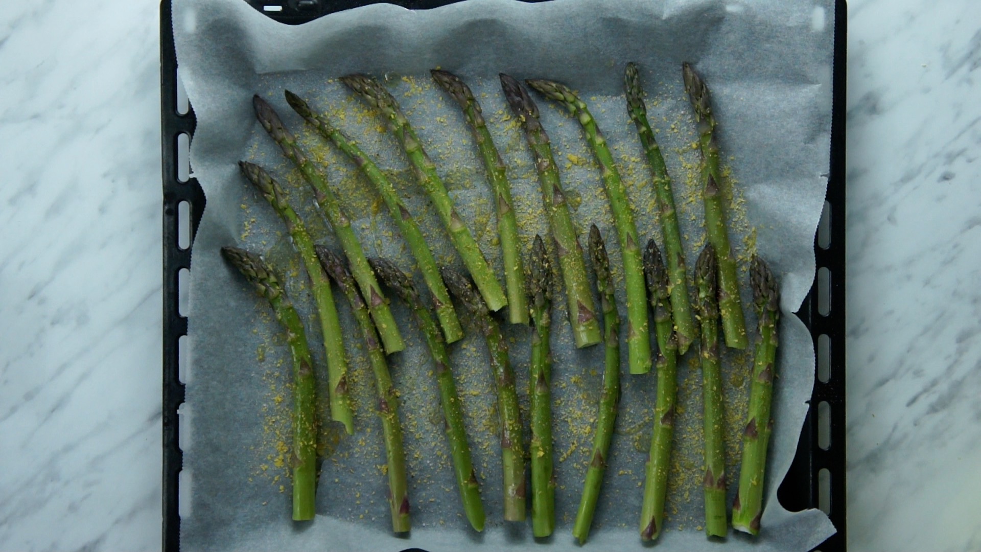 Green asparagus on a baking sheet and parchment paper sprinkled with salt, black pepper, and yellow flakes.
