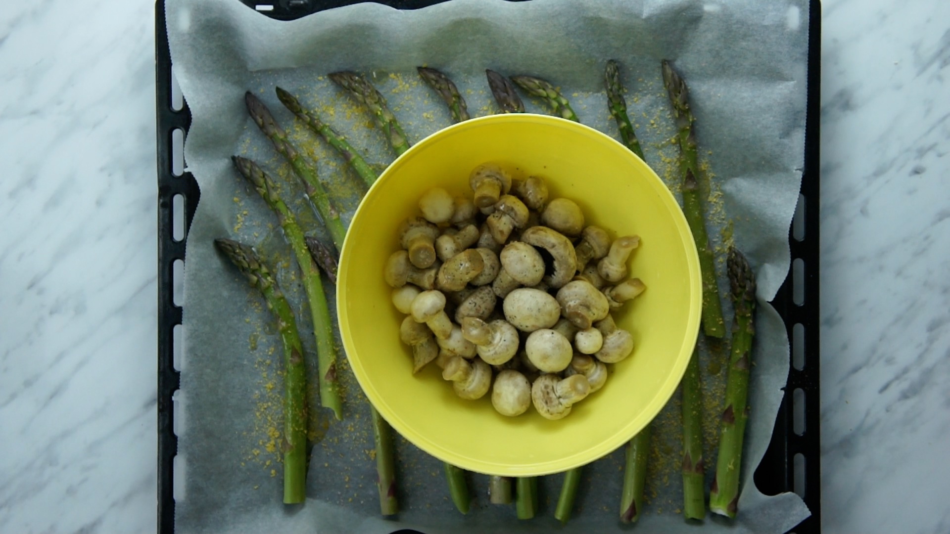 Green asparagus on a baking sheet and parchment paper sprinkled with salt, black pepper, and yellow flakes. There is a yellow plastic bowl on top with small mushrooms.