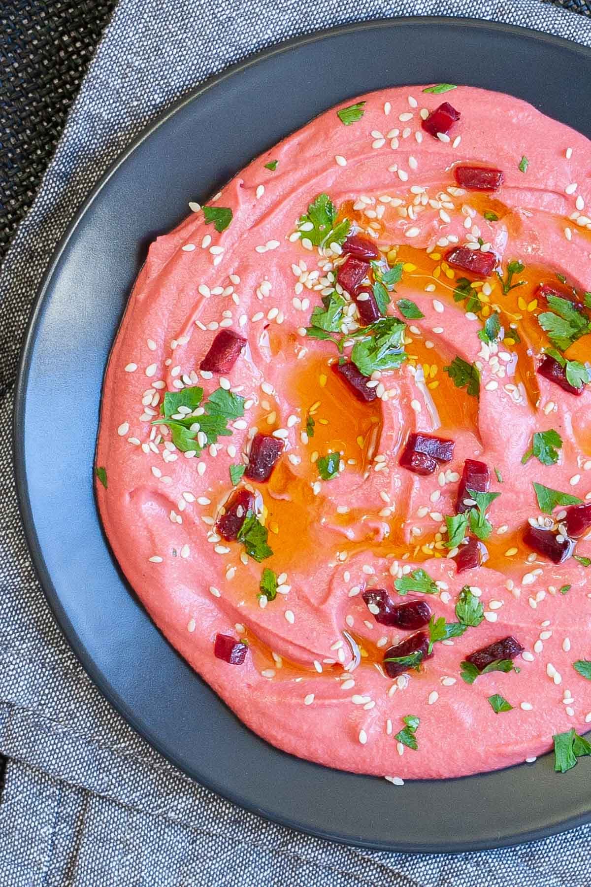 Pink hummus on a black plate, sprinkled with freshly chopped parsley, sesame seeds and small roasted beet pieces.