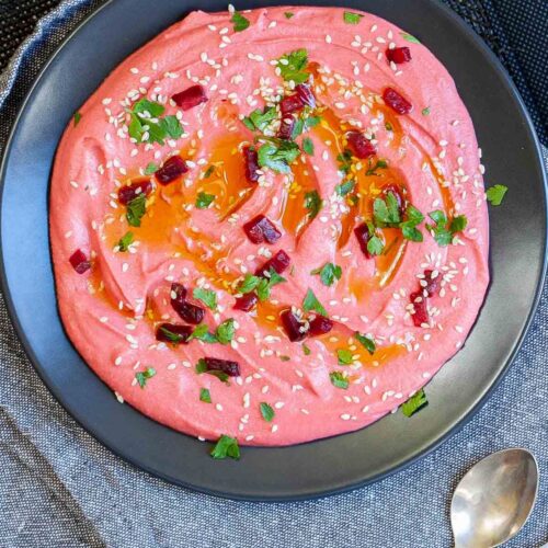 Pink hummus on a black plate, sprinkled with freshly chopped parsley, sesame seeds and small roasted beet pieces.