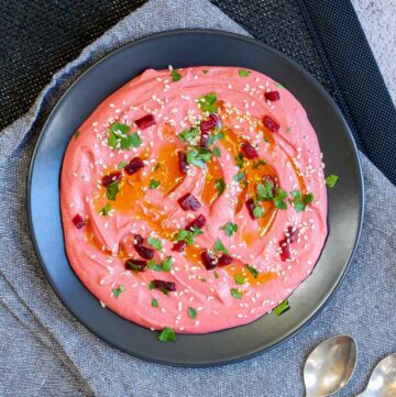 Pink hummus on a black plate, sprinkled with freshly chopped parsley, sesame seeds and small roasted beet pieces.