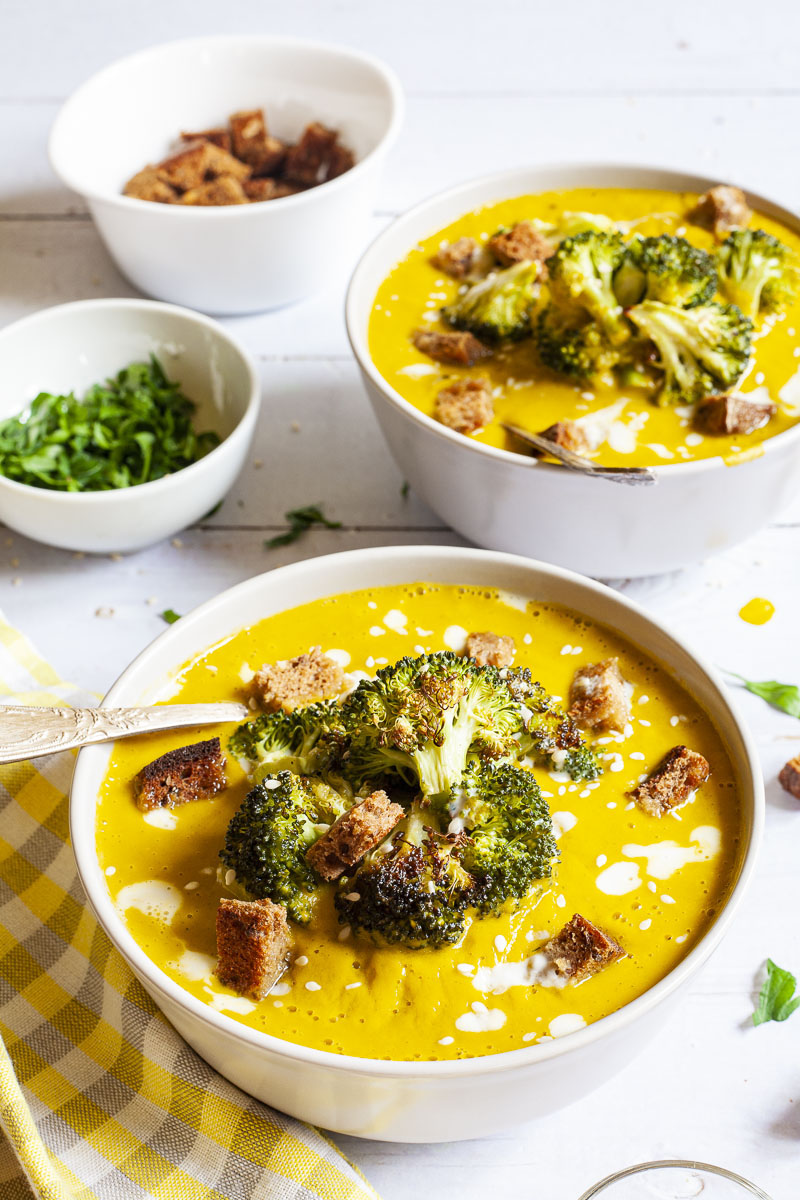 Yellow cream soup topped with roasted broccoli florets, brown croutons, white drizzle spots served in 2 white bowls with a spoon in each. Other toppings like croutons, red pepper flakes and chopped parsley is in small bowls around the soups.