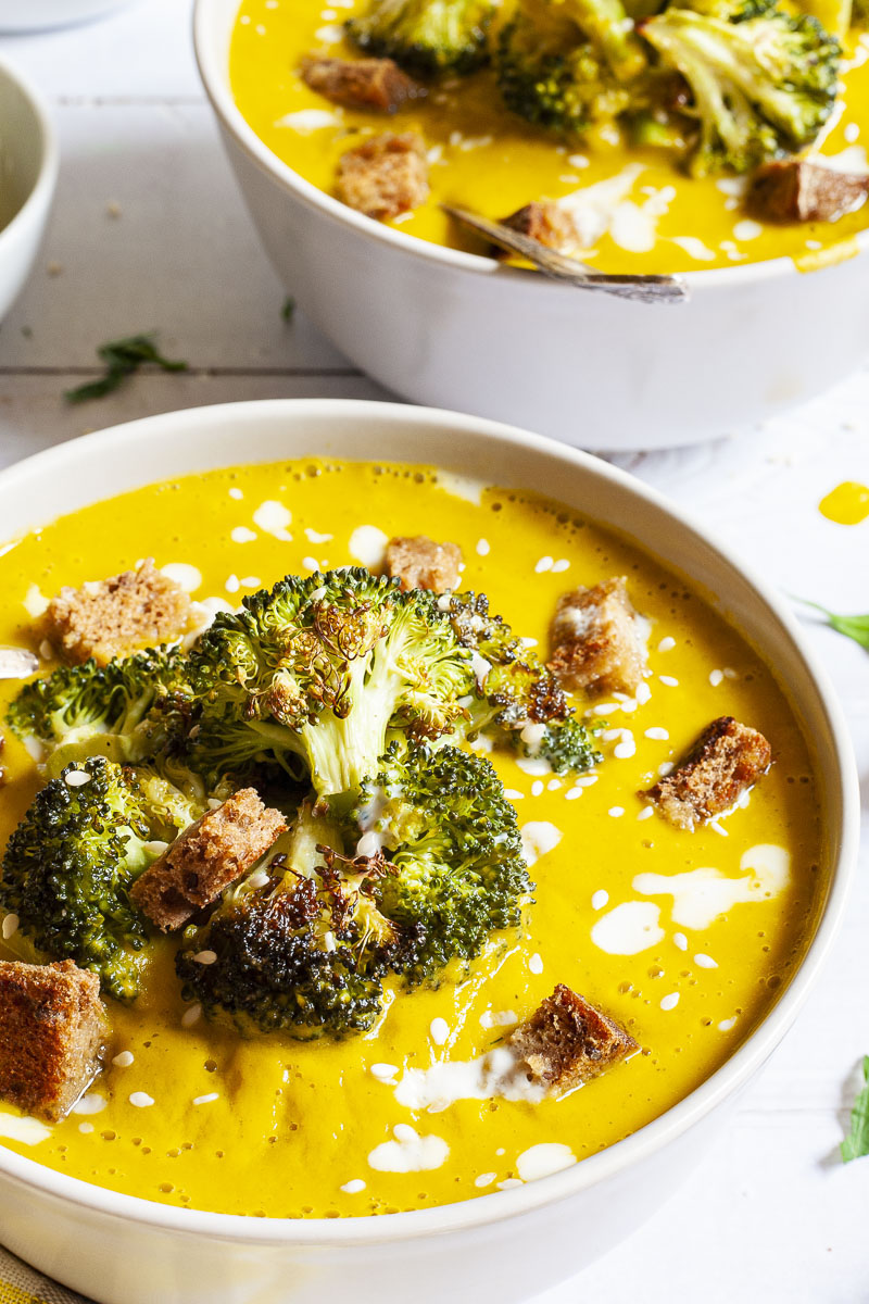 Yellow cream soup topped with roasted broccoli florets, brown croutons, white drizzle spots served in 2 white bowls with a spoon in each. Other toppings like croutons, red pepper flakes and chopped parsley is in small bowls around the soups.