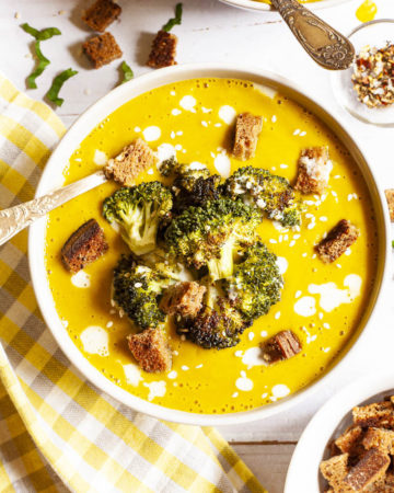 Yellow cream soup topped with roasted broccoli florets, brown croutons, white drizzle spots served in 2 white bowls with a spoon in each. Other toppings like croutons, red pepper flakes and chopped parsley is in small bowls around the soups.