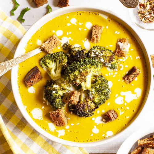 Yellow cream soup topped with roasted broccoli florets, brown croutons, white drizzle spots served in 2 white bowls with a spoon in each. Other toppings like croutons, red pepper flakes and chopped parsley is in small bowls around the soups.