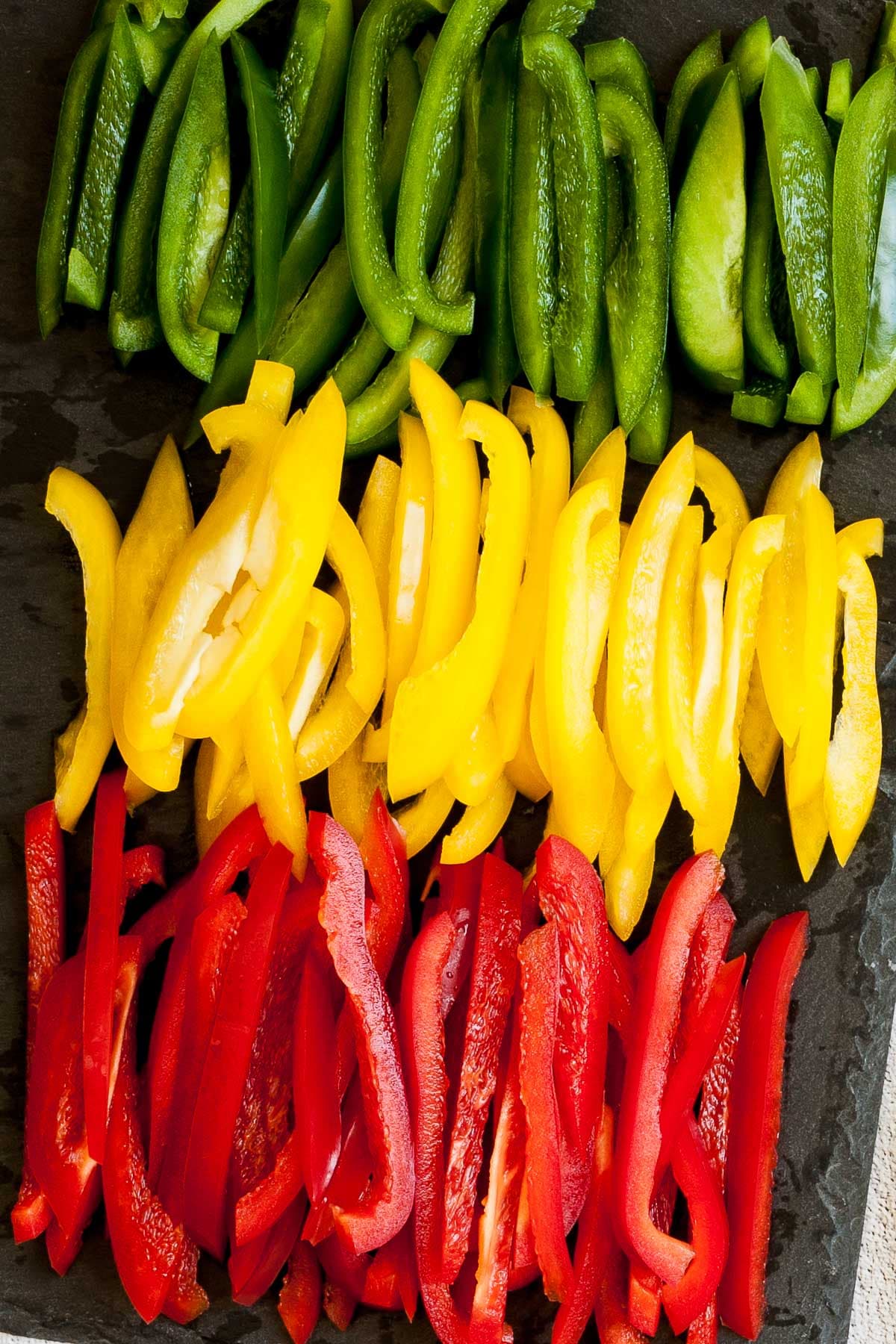 Raw red yellow and green bell pepper slices nicely stakced next to each other on a black board.