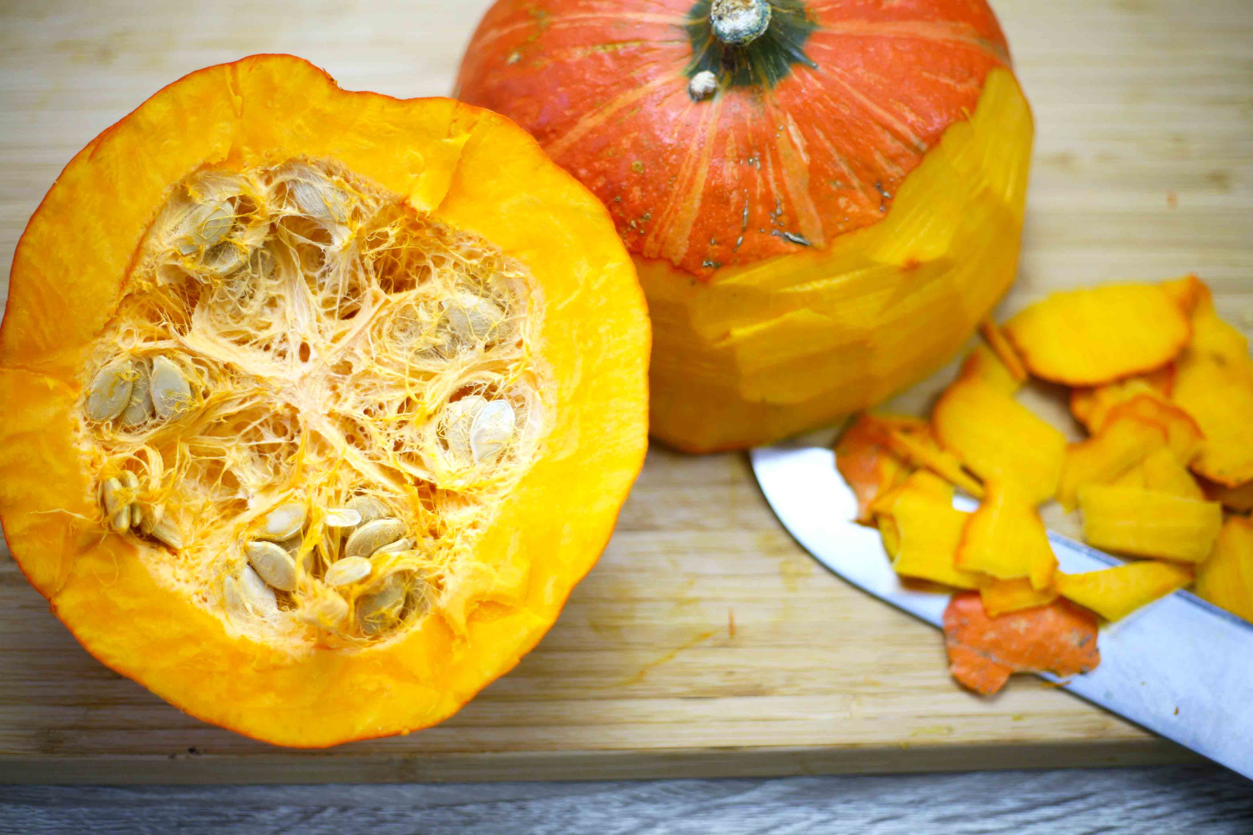 An orange squash is cut in halve and you can see the seeds in the middle. The other half is turned upside down and a knife is carving off the skin
