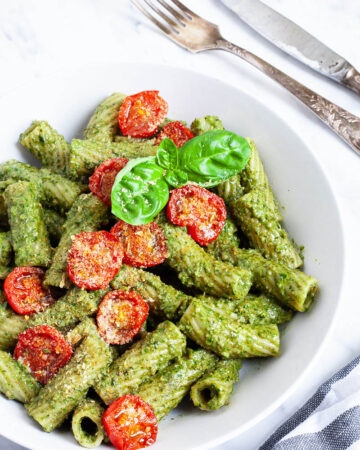 White bowl with tortiglioni pasta covered in green pesto topped with cherry tomato halves and sprinkled with yellow flakes.