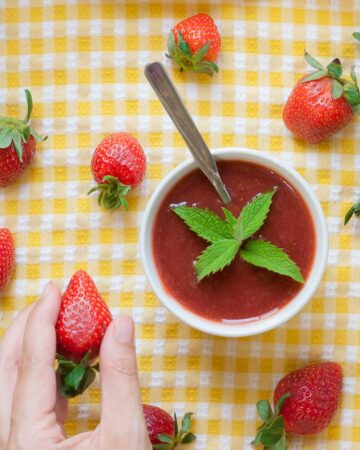 Two small white bowls with strawberry puree and some mint leaves. More fresh strawberries are scattered around it on a yellow tablecloth. A hand is holding one strawberry.