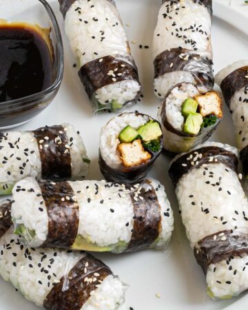 Black and white rice paper rolls on a large white plate sprinkled with black and white sesame seeds. One is cut in the middle so the filling of tofu sticks, cucumber, avocado, and chives is visible.