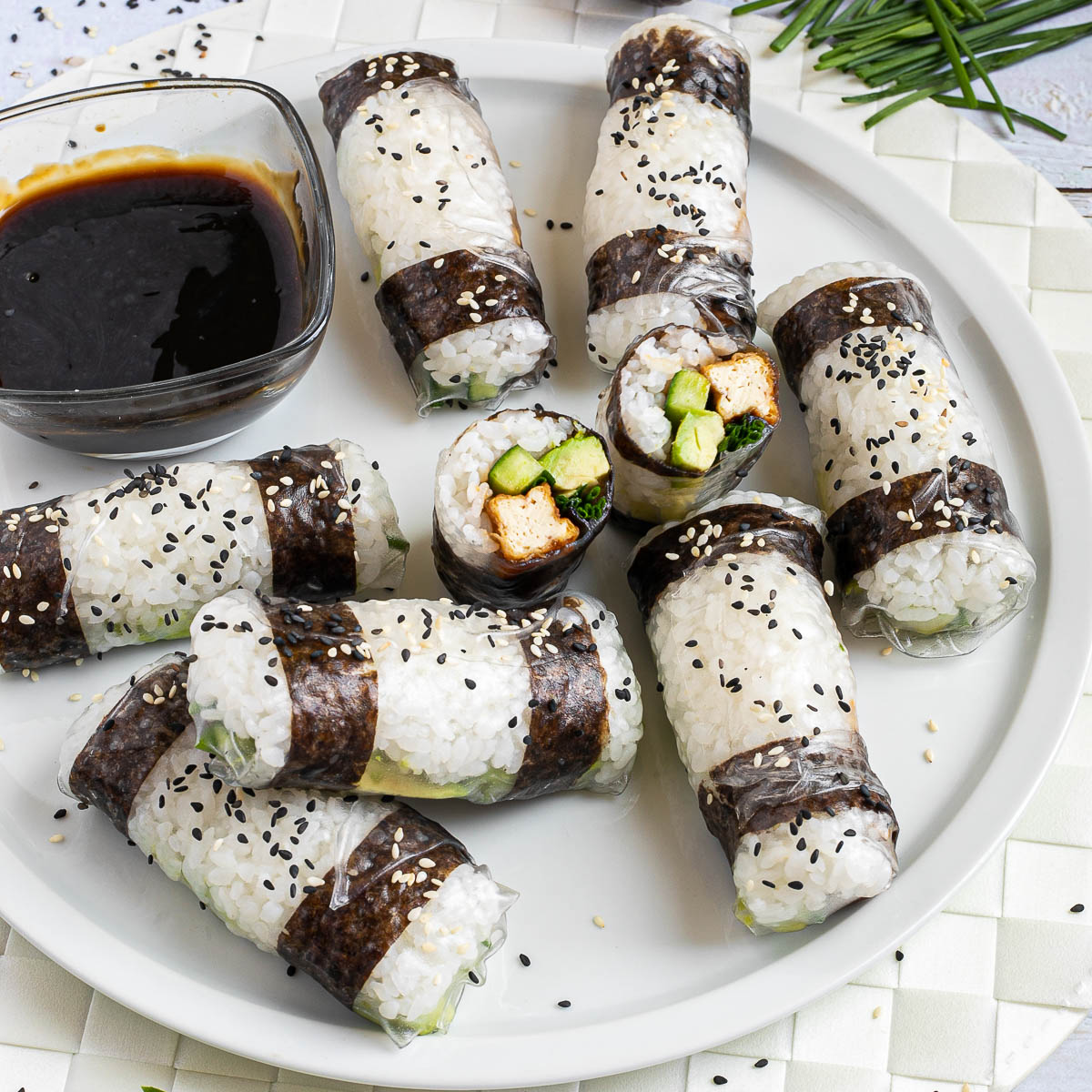 Black and white rice paper rolls on a large white plate sprinkled with black and white sesame seeds. One is cut in the middle so the filling of tofu sticks, cucumber, avocado, and chives is visible.