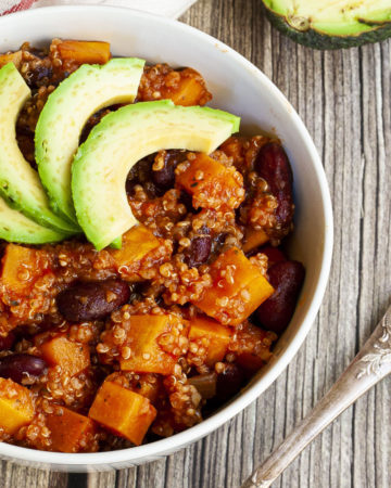 In a small white bowl orange cubes, dark brown beans, small white, yellow seeds with 3 slices of green avocado on top.