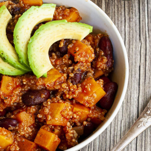 In a small white bowl orange cubes, dark brown beans, small white, yellow seeds with 3 slices of green avocado on top.