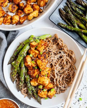 Large white bowl with brown soba noodles, orange crispy tofu bites, roasted asparagus and black and white sesame seeds. More asparagus and tofu bites are on the side. Chopsticks are placed on the side.