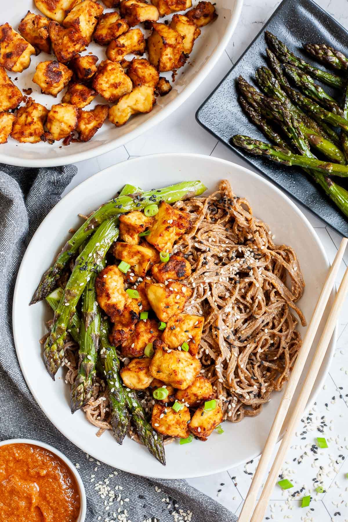 Large white bowl with brown soba noodles, orange crispy tofu bites, roasted asparagus and black and white sesame seeds. More asparagus and tofu bites are on the side. Chopsticks are placed on the side.