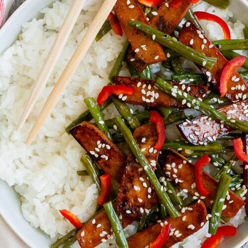 White bowl from above with white rice topped with thin dark brown tofu slices with charred edges, green beans, chopped red bell pepper and sprinkled with white sesame seeds. Light brown chopsticks are placed on the side.