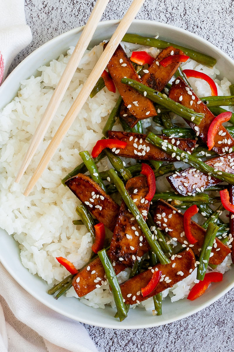 White bowl from above with white rice topped with thin dark brown tofu slices with charred edges, green beans, chopped red bell pepper and sprinkled with white sesame seeds. Light brown chopsticks are placed on the side.