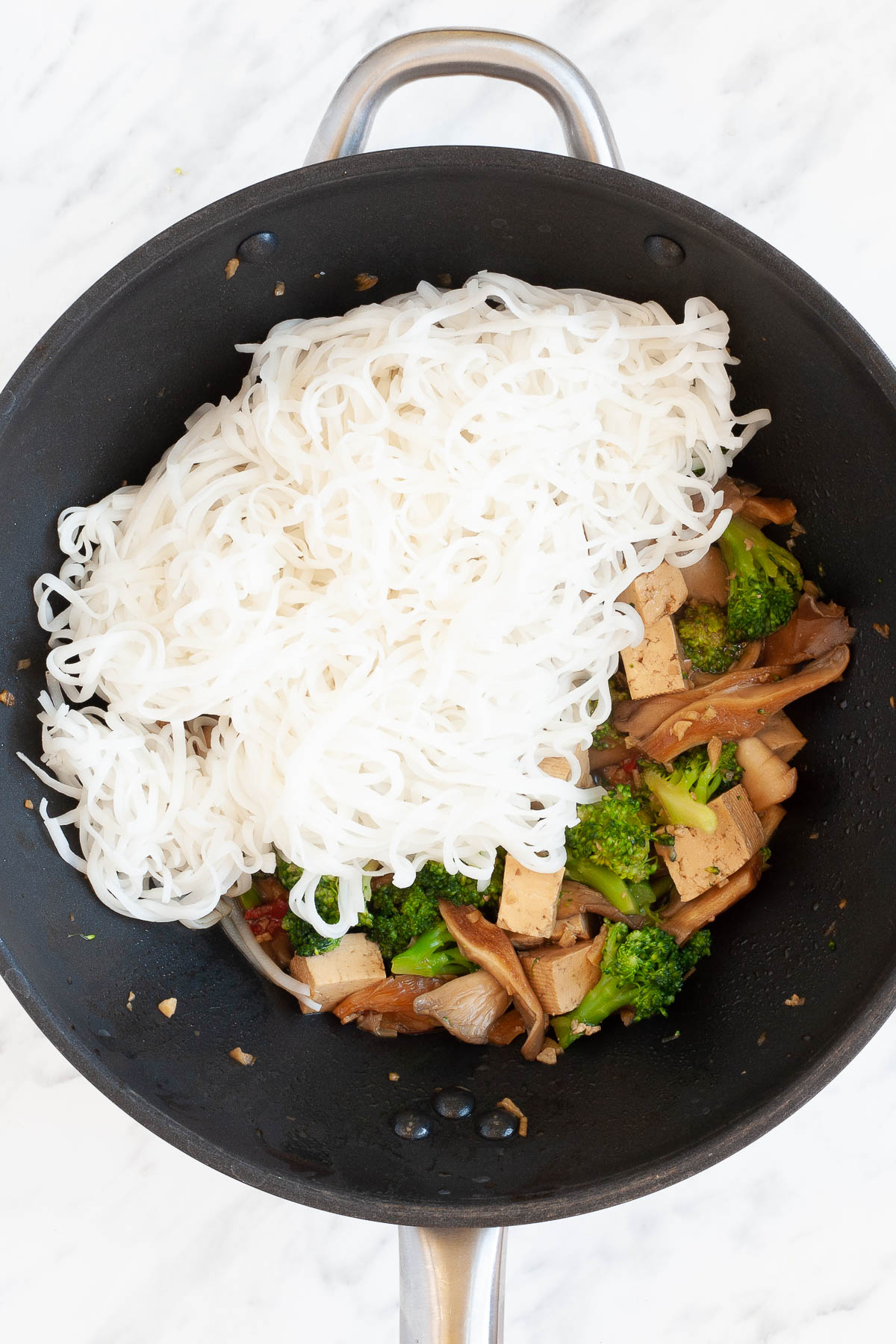 Black wok with mushroom shreds, tofu cubes, broccoli florets brown due to a bit of sauce topped with a heap of rice noodles.