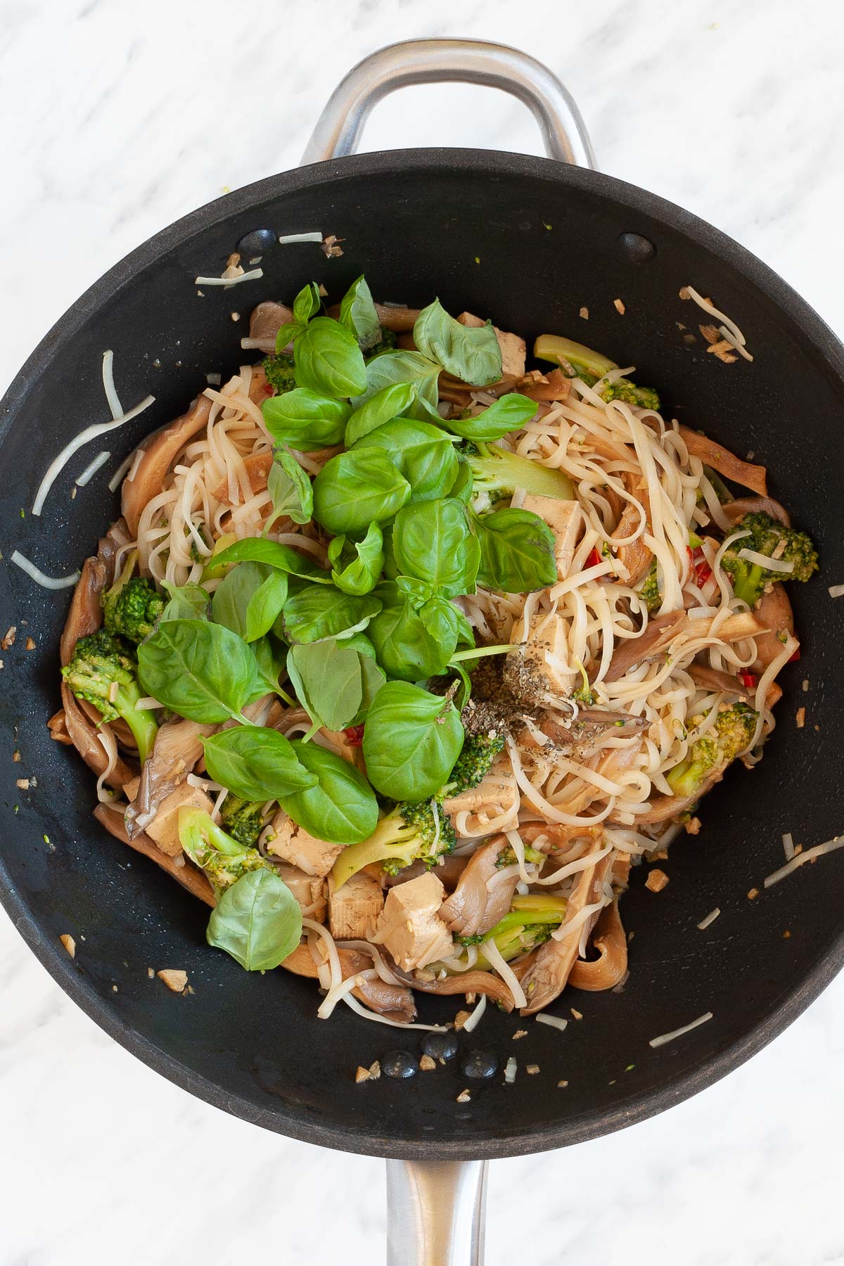 Black wok with noodles, mushroom shreds, tofu cubes, broccoli florets topped with fresh herbs.