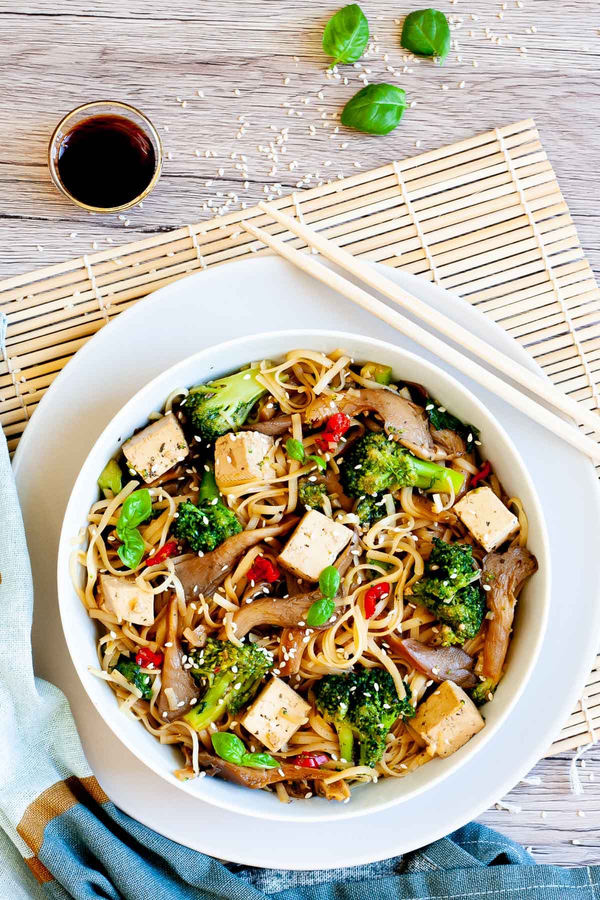 White bowl with tofu cubes, broccoli florets, oyster mushroom shreds, red chili slices and long thin rice noodles.