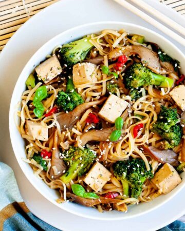 White bowl with tofu cubes, broccoli florets, oyster mushroom shreds, red chili slices and long thin rice noodles.