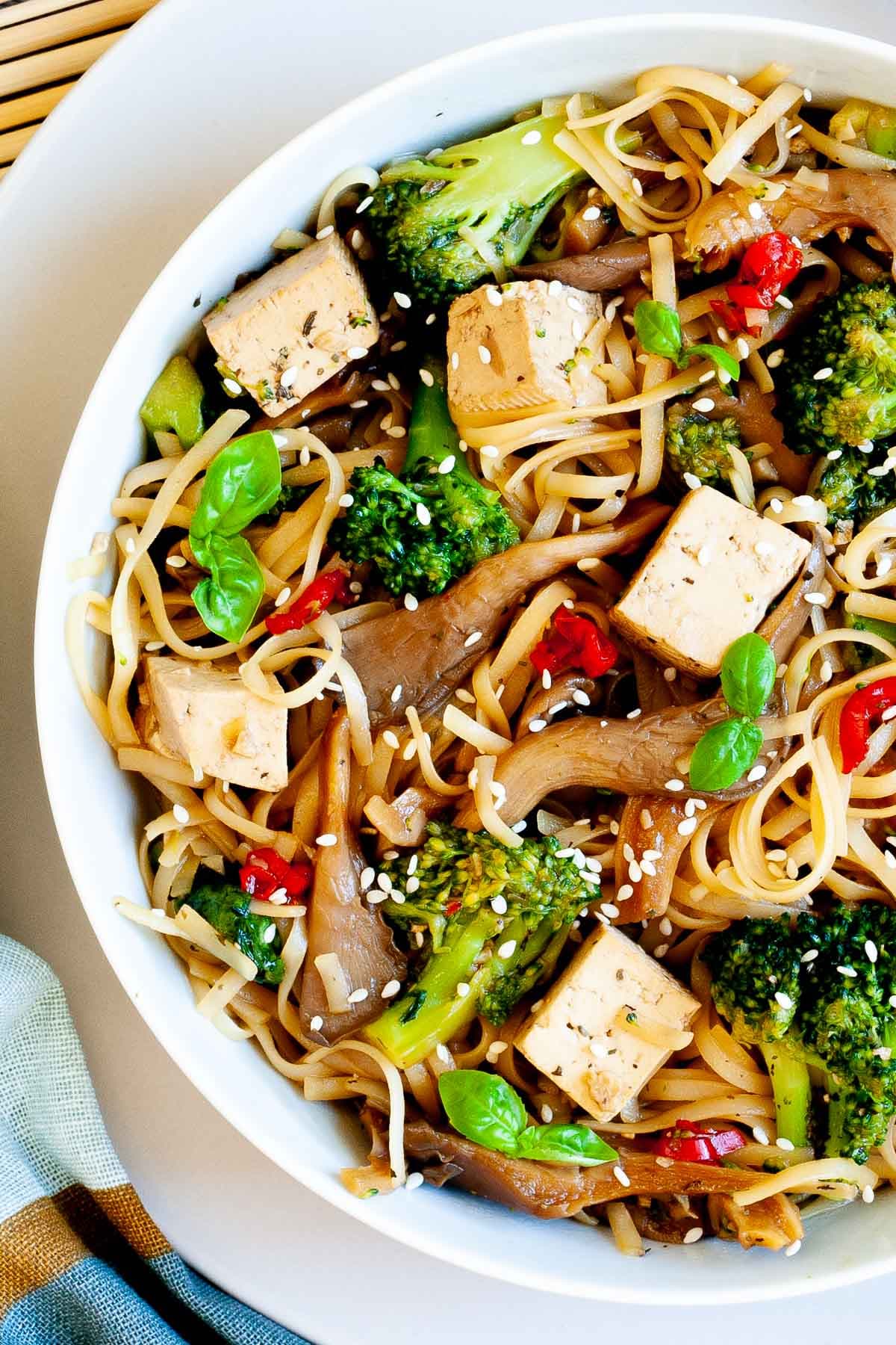 White bowl with tofu cubes, broccoli florets, oyster mushroom shreds, red chili slices and long thin rice noodles.