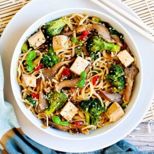 White bowl with tofu cubes, broccoli florets, oyster mushroom shreds, red chili slices and long thin rice noodles.