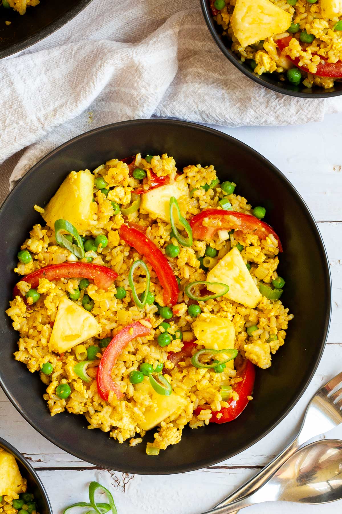 A black bowl with yellow rice, red tomato slices, pineapple chunks, green peas and chopped green onion. A skillet with the leftovers is in the corner.