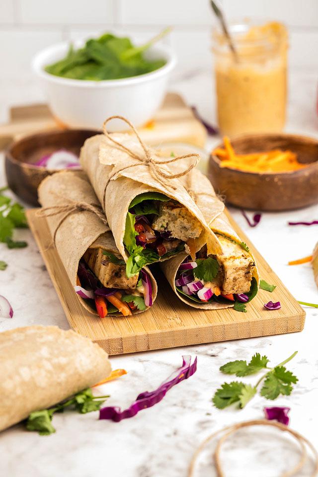 3 wraps with colorful veggies and tofu slices are placed on top of each other on a cutting board.