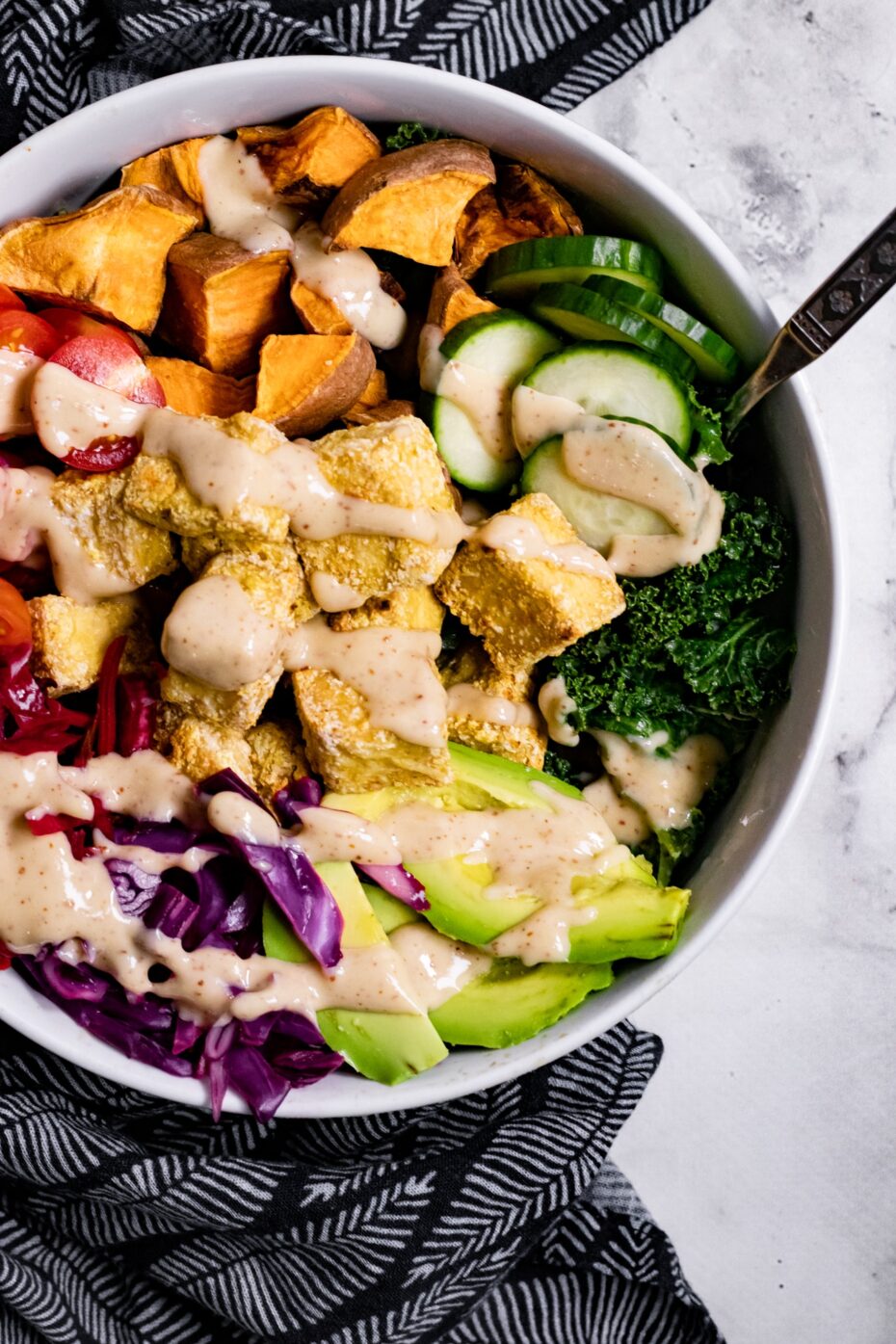 White bowl from above with avocado slices, cucumber, purple cabbage, tofu cubes and diced sweet potato drizzled with a light brown dressing