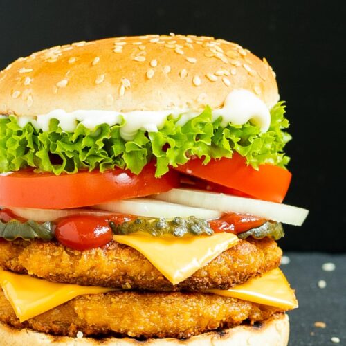 Burger bun with two patties, cheese in between, pickled cucumber slices, onion slices, tomato and ruffled lettuce.