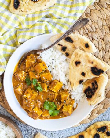 A white plate with white rice, pieces of naan bread and tofu cubes in a creamy orange sauce sprinkled with sesame seeds and freshly chopped cilantro. A spoon is placed inside.