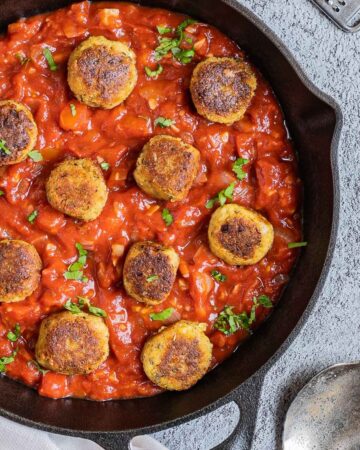 Black cast iron skillet with red marinara sauce and crispy brown tofu meatballs. It is sprinkled with chopped fresh green herbs.