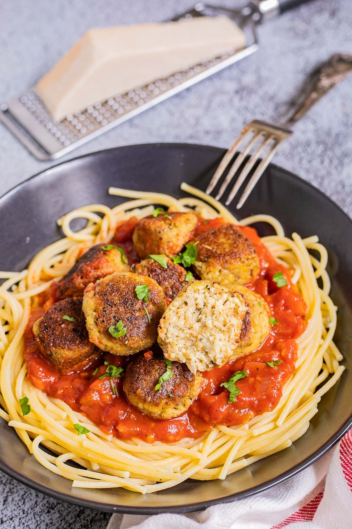 Black plate with spaghetti, marinara sauce and crispy brown tofu meatballs sprinkled with chopped green herbs. One meatballs is cut in half so you can see the inside. A fork is placed next to it and vegan parmesan cheese on top of a cheese grater.