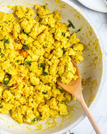 Yellow tofu crumbles in a white frying pan sprinkled with fresh herbs and red pepper flakes. A wooden spatula is placed inside.