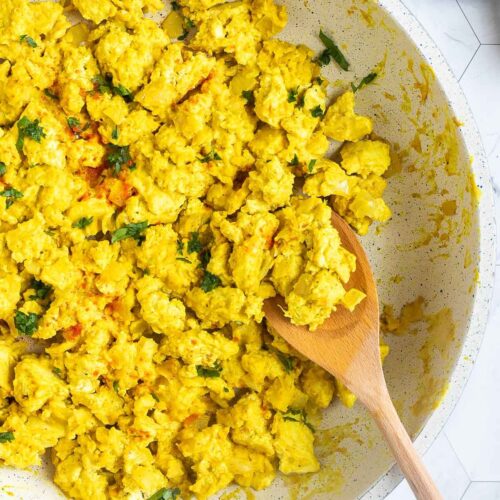 Yellow tofu crumbles in a white frying pan sprinkled with fresh herbs and red pepper flakes. A wooden spatula is placed inside.