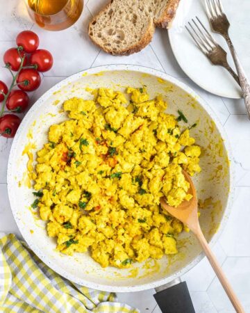 Yellow tofu crumbles in a white frying pan sprinkled with fresh herbs and red pepper flakes. A wooden spatula is placed inside.