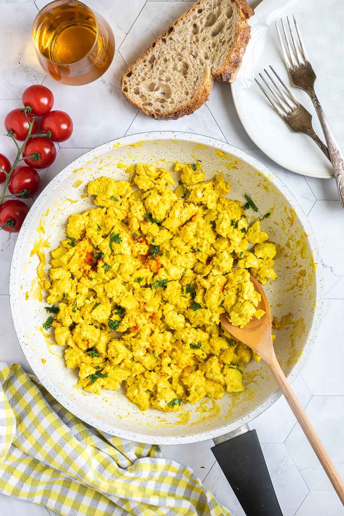Yellow tofu crumbles in a white frying pan sprinkled with fresh herbs and red pepper flakes. A wooden spatula is placed inside.