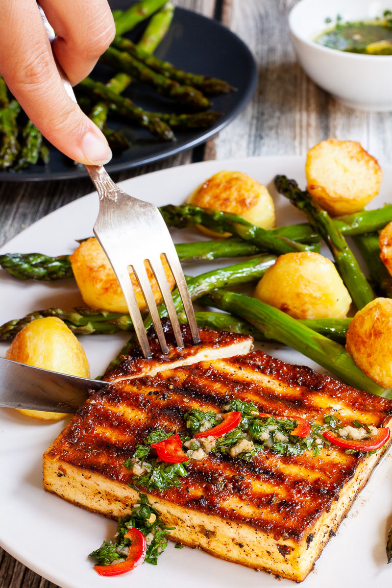 Light grey round plate with a brown, shiny tofu slice topped with a chopped green herbs served with baked half potatoes and roasted asparagus. A hand is using a fork and a knife to cut off the corner.
