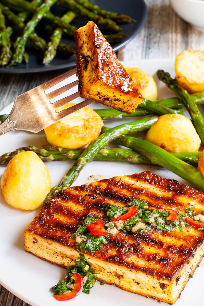Light grey round plate with a brown, shiny tofu slice topped with a chopped green herbs served with baked half potatoes and roasted asparagus. A hand is holding a fork with one bite.
