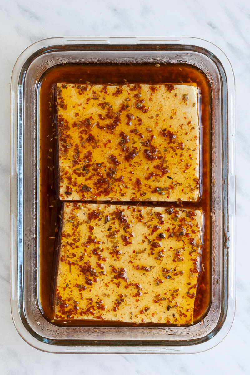 A glass bowl with two tofu slices swimming in a oily brown sauce with dried herbs.