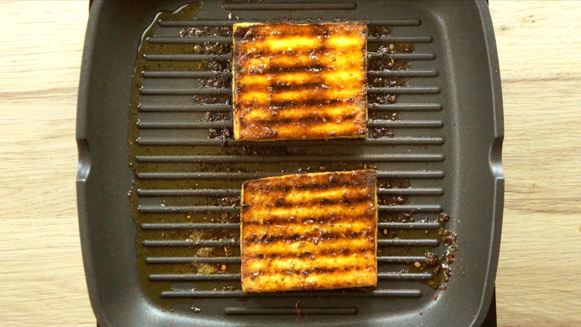 Two square brown tofu slices are charcoaled in a grill pan