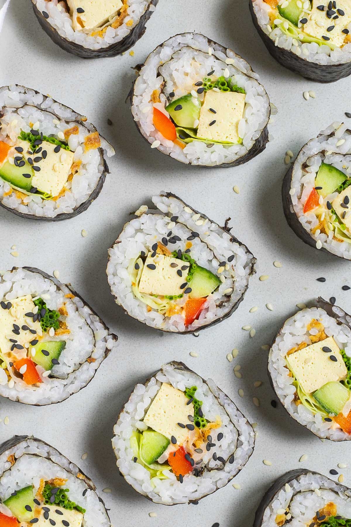 An oval, light blue plate with 9 tofu sushi rolls with colorful filling wrapped in dark seaweed sheets.