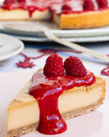 One slice of cheesecake with vibrant red sauce and raspberries on top is served on a light pink plate. The whole cake is in the background.