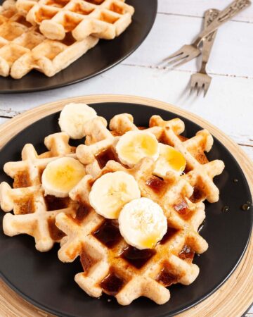 Black plate with a stack of waffles topped with sliced banana and maple syrup. There are 3 waffles on another black plate in the background.