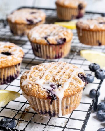 Several lemon muffins with blueberries on a black wire rack drizzled with a white glaze. Lemon slices and fresh blueberries are scattered around them.